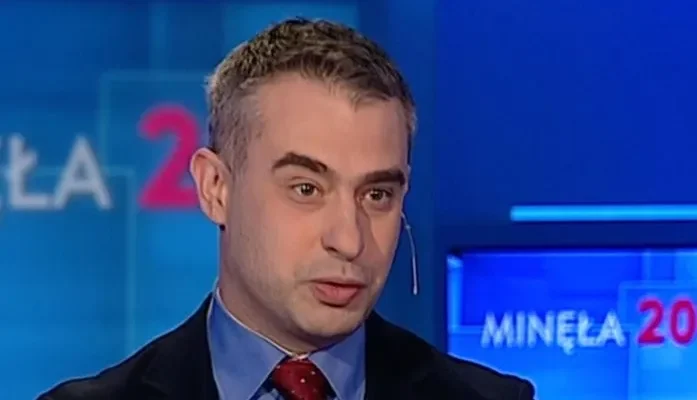 Mężczyzna w garniturze i czerwonym krawacie z mikrofonem nasadnym, mówiący w studiu telewizyjnym na niebieskim tle z widocznym napisem programu.