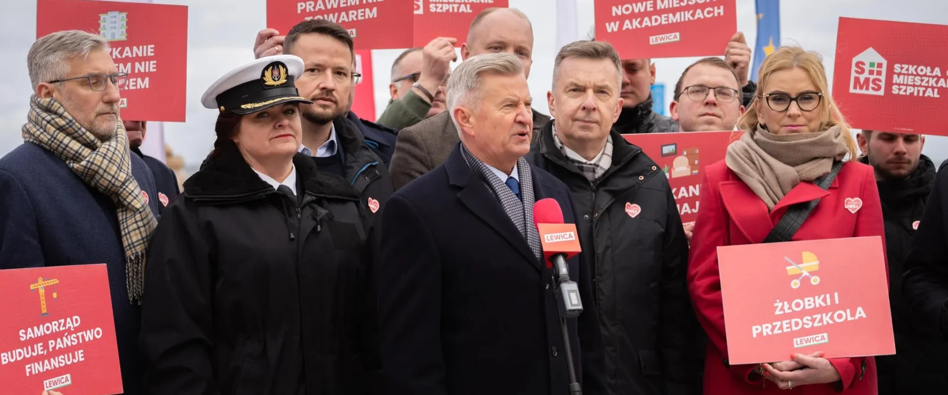 Grupa działaczy Lewicy podczas wiecu, na pierwszym planie mężczyzna w garniturze przemawia do mikrofonu, wokół niego stoją osoby z czerwonymi plakatami promującymi inwestycje w szkoły, mieszkania, żłobki i inne usługi.