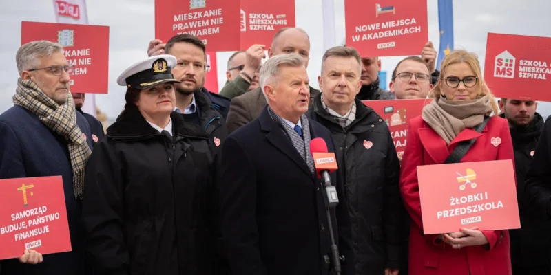 Grupa działaczy Lewicy podczas wiecu, na pierwszym planie mężczyzna w garniturze przemawia do mikrofonu, wokół niego stoją osoby z czerwonymi plakatami promującymi inwestycje w szkoły, mieszkania, żłobki i inne usługi.