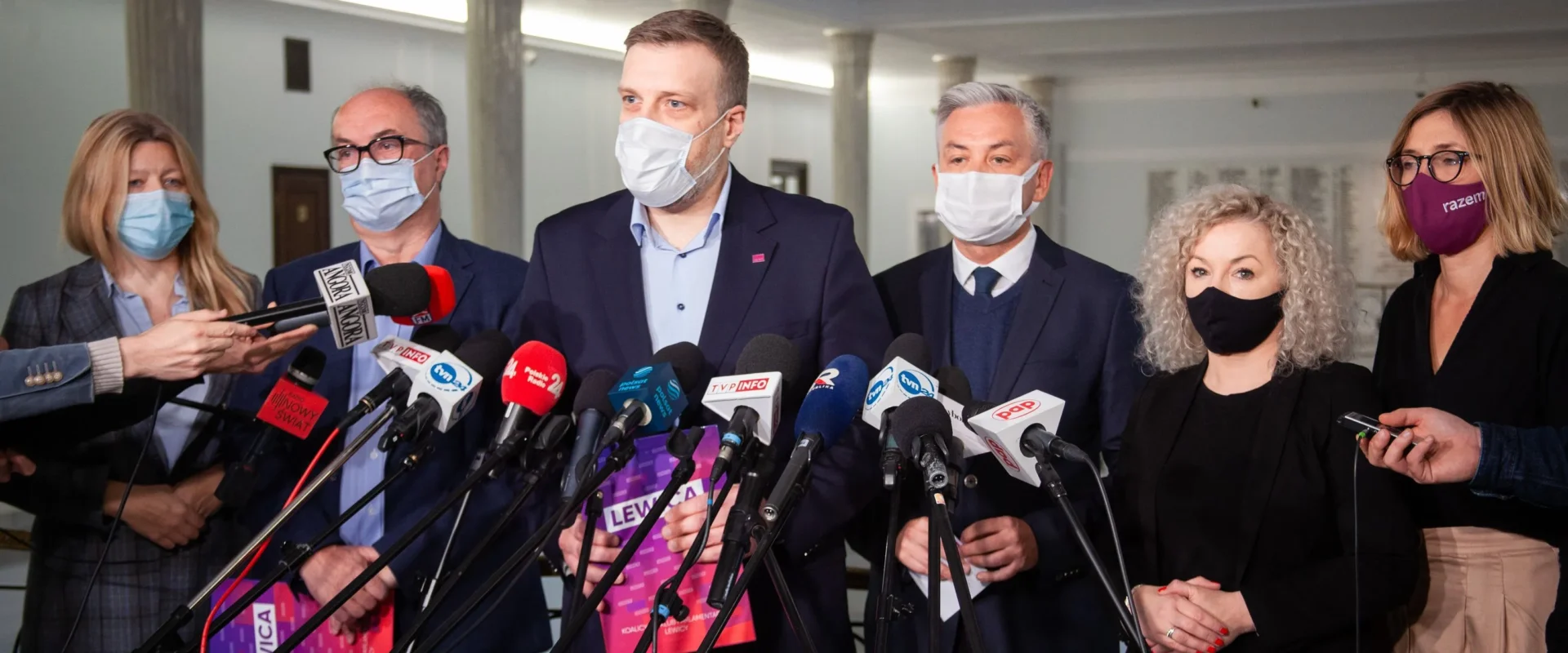 Konferencja prasowa, na której obecni są politycy w maskach ochronnych, otoczeni mikrofonami w sali z jasnym oświetleniem.