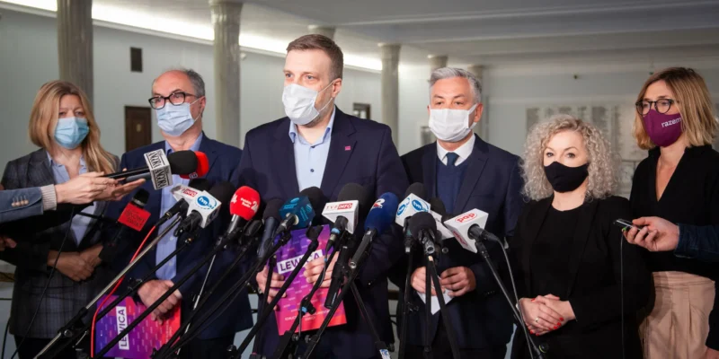 Konferencja prasowa, na której obecni są politycy w maskach ochronnych, otoczeni mikrofonami w sali z jasnym oświetleniem.