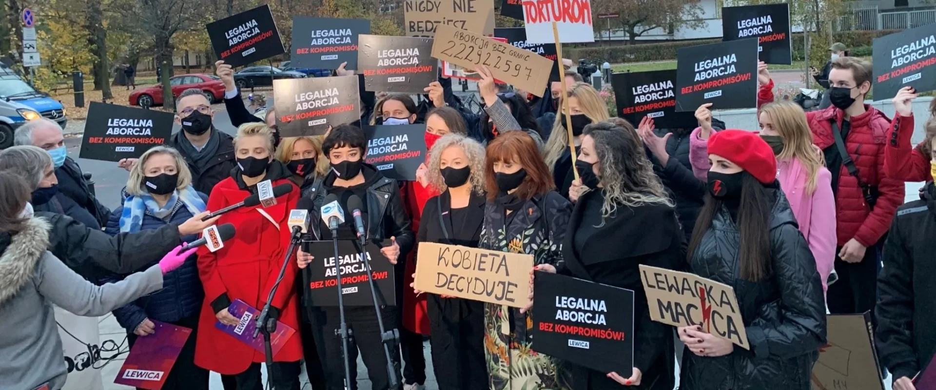 Grupa demonstrantów Lewicy z plakatami „Legalna aborcja bez kompromisów” i hasłem „Kobieta decyduje”, stojąca przed mikrofonami mediów podczas protestu.