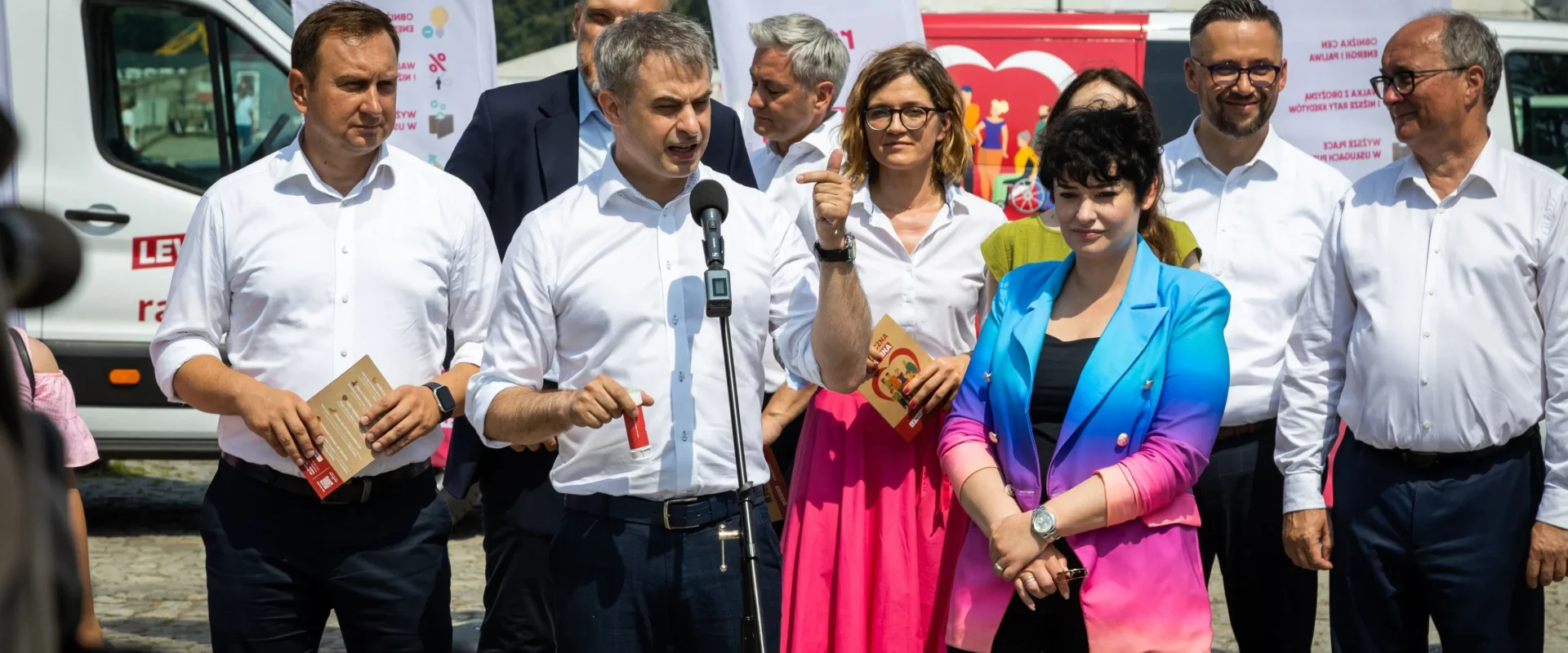 Grupa polityków Lewicy w białych koszulach stoi na zewnątrz podczas obchodów Święta Pracy; centralny mężczyzna przemawia do mikrofonu, a w tle widać banery Lewica i białą furgonetkę z napisem LEWICA.