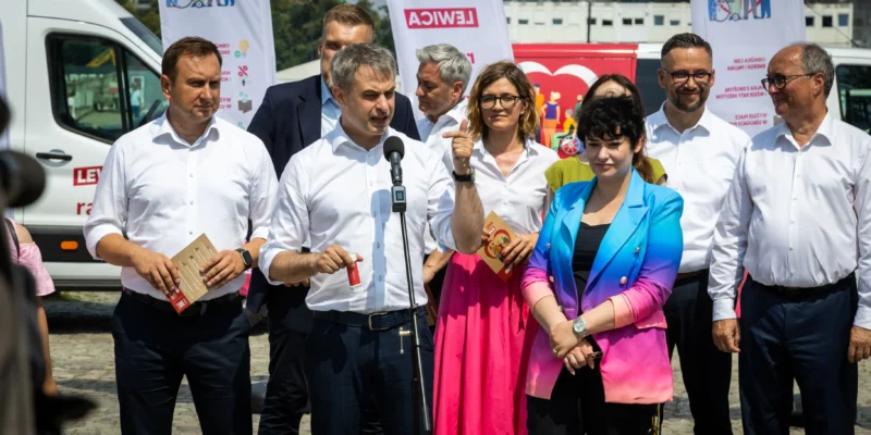 Grupa polityków Lewicy w białych koszulach stoi na zewnątrz podczas obchodów Święta Pracy; centralny mężczyzna przemawia do mikrofonu, a w tle widać banery Lewica i białą furgonetkę z napisem LEWICA.