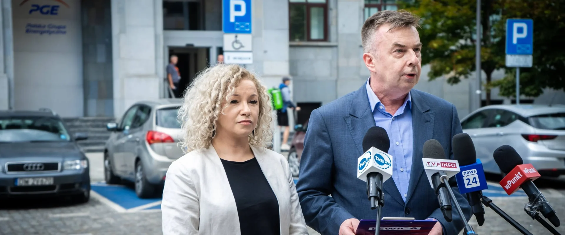 Mężczyzna w garniturze przemawia do zestawu mikrofonów dziennikarskich stojąc przed wejściem do budynku PGE GiEK, obok stoi kobieta.