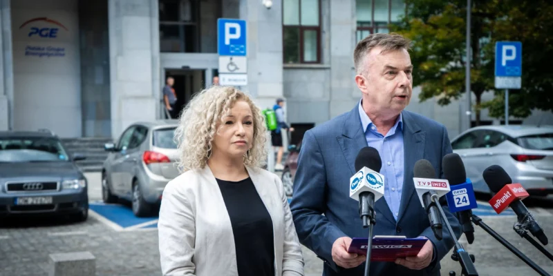 Mężczyzna w garniturze przemawia do zestawu mikrofonów dziennikarskich stojąc przed wejściem do budynku PGE GiEK, obok stoi kobieta.