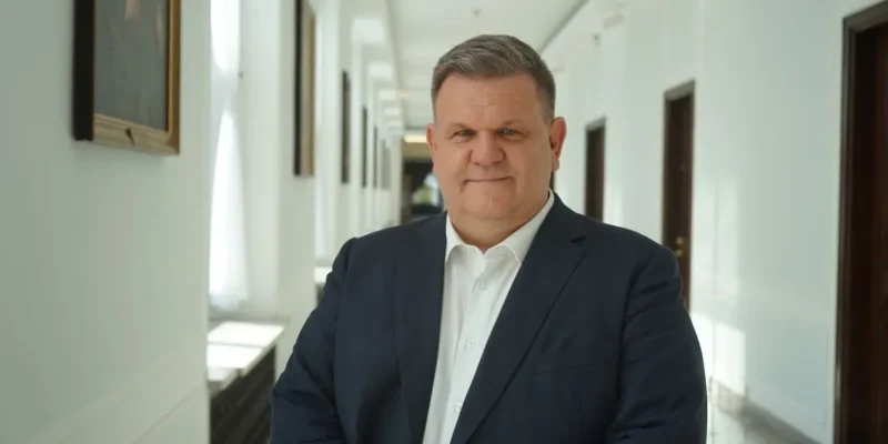 Mężczyzna w granatowym garniturze i białej koszuli stoi w jasnym korytarzu z obrazami na ścianach, patrząc przed siebie.