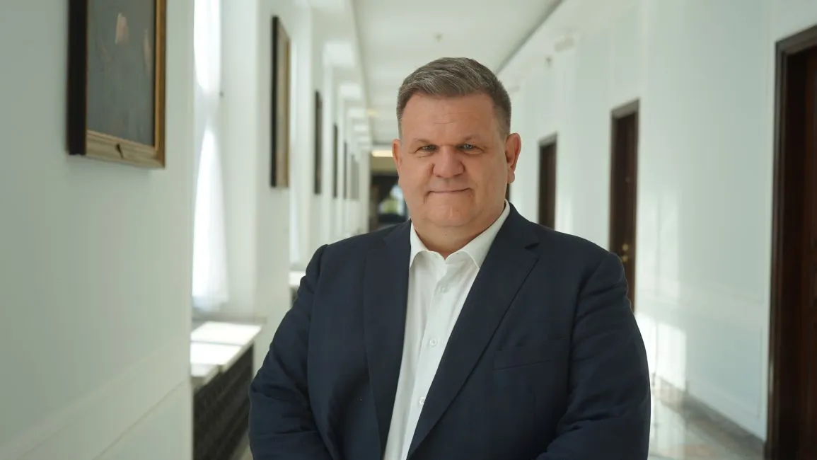 Mężczyzna w granatowym garniturze i białej koszuli stoi w jasnym korytarzu z obrazami na ścianach, patrząc przed siebie.