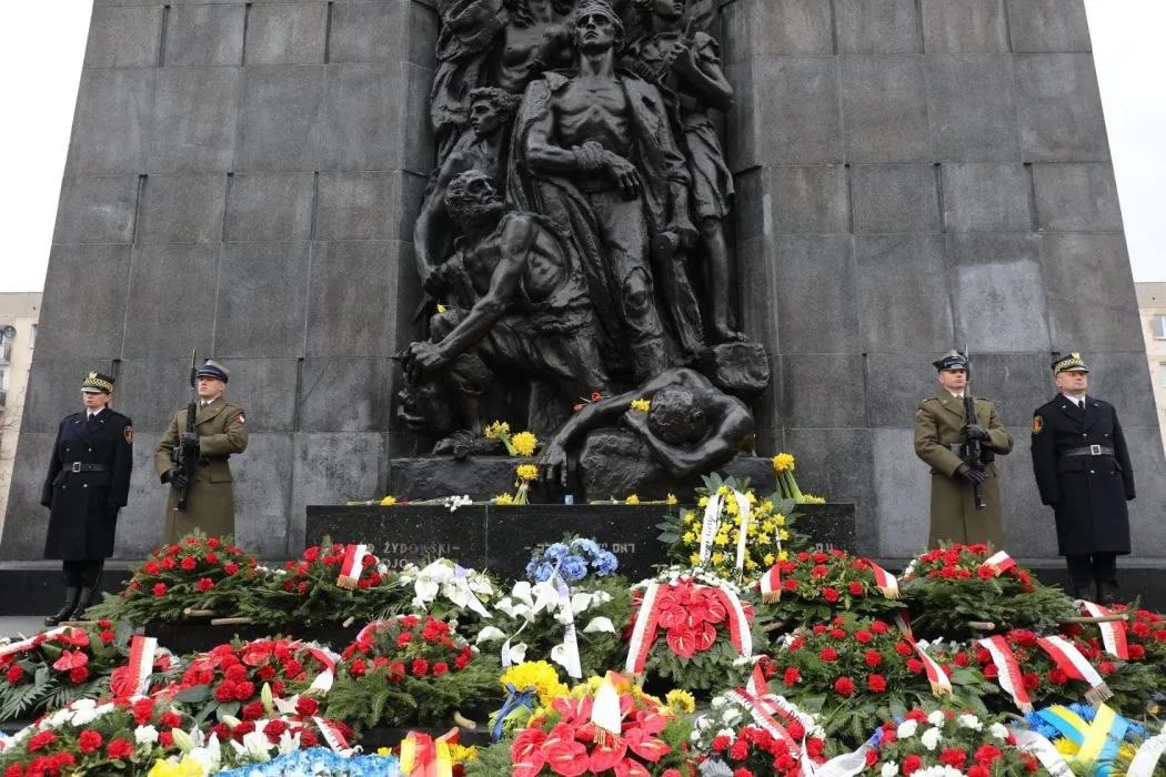 Żołnierze w uniformach stoją na straży przed monumentalnym pomnikiem z rzeźbą upamiętniającym Powstanie w Getcie Warszawskim, wokół którego leżą liczne wieńce i kwiaty.
