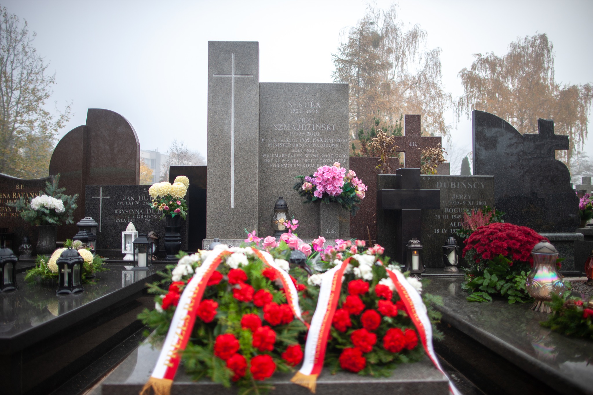 Na zdjęciu widoczny jest cmentarz z kilkoma nagrobkami, ozdobionymi kwiatami i świeczkami, w mglistej atmosferze.