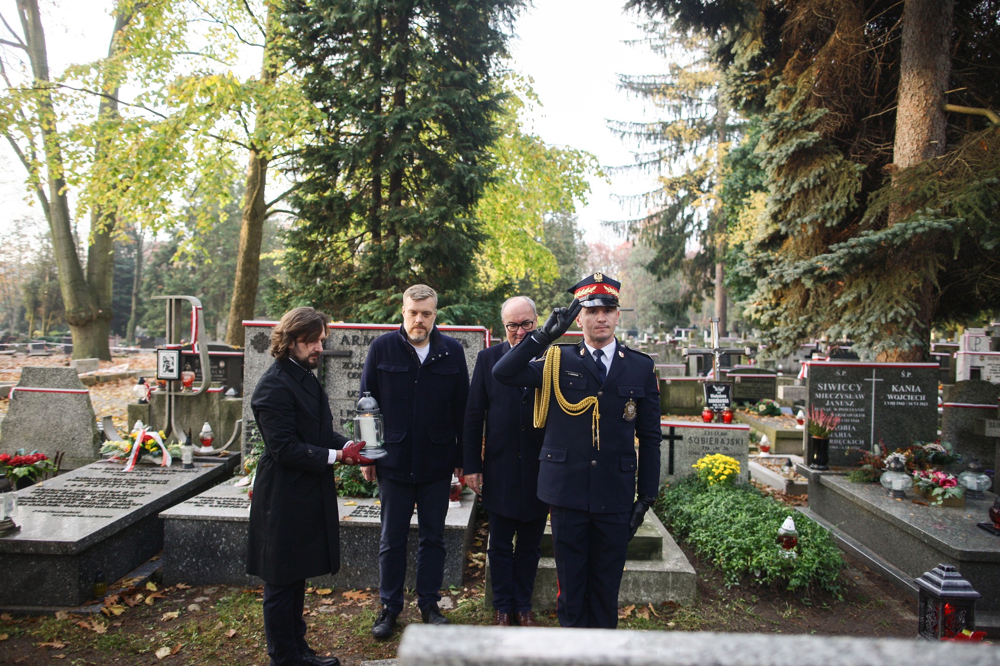 Na zdjęciu widoczni są mężczyźni na cmentarzu, z jednym w mundurze wojskowym, wykonującym salut, podczas gdy inni trzymają znicz.