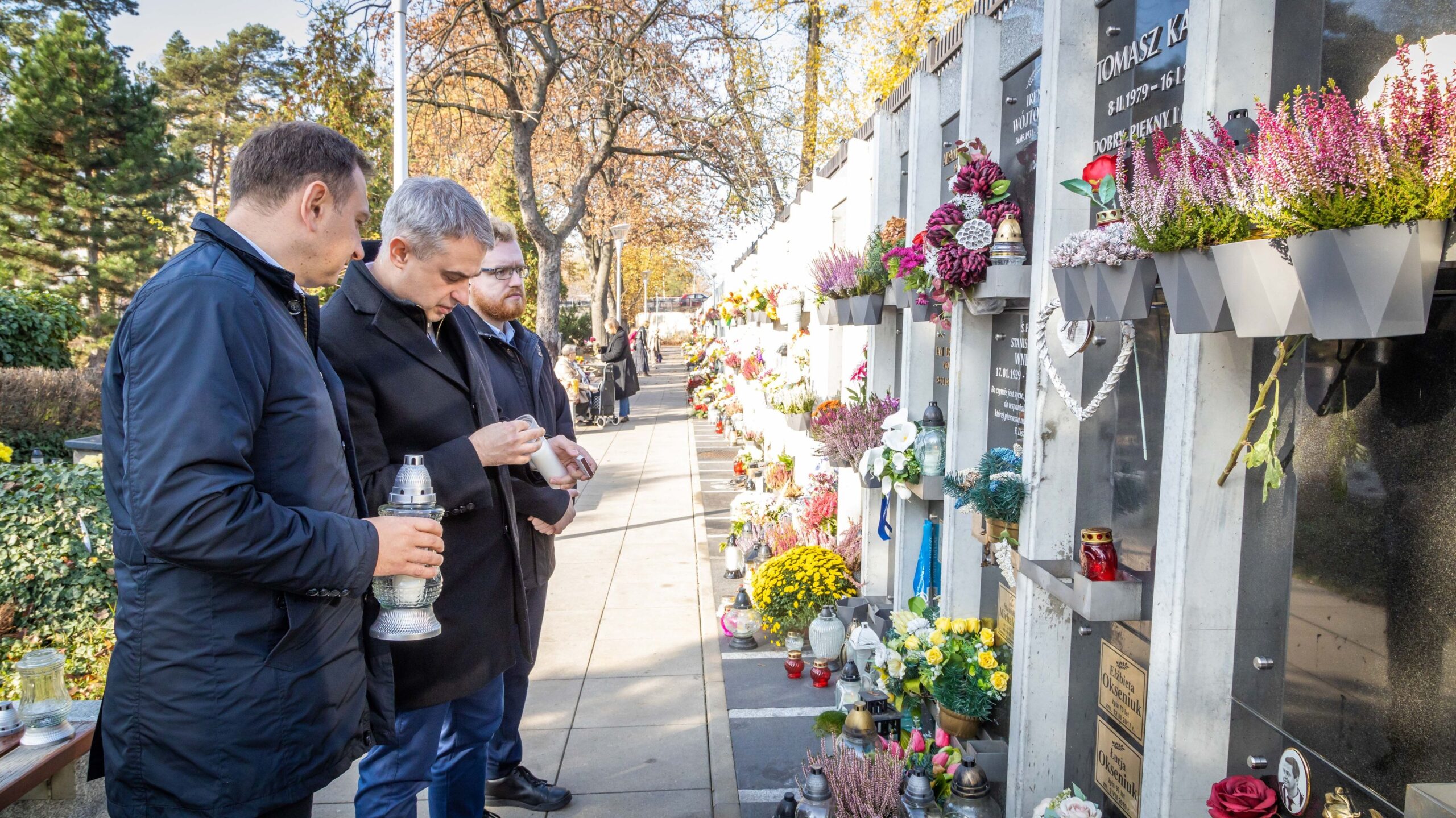 Trzej mężczyźni odwiedzają cmentarz, gdzie z zapalonymi zniczami stoją przy grobach, otoczonych kwiatami i dekoracjami.