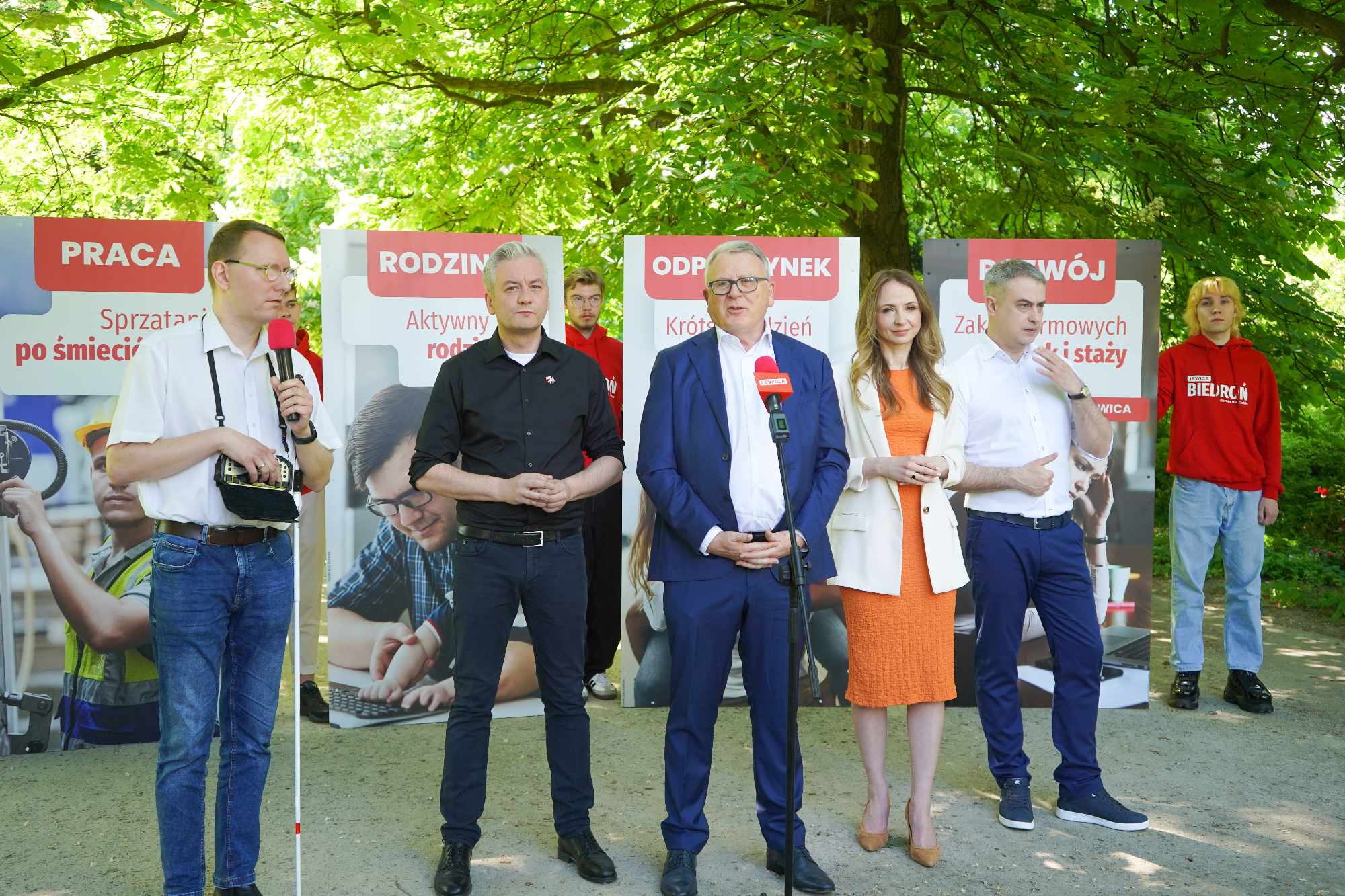 Grupa osób na zewnątrz podczas Święta Pracy 2024, prowadzących przemówienie przed planszami z hasłami o pracy, rodzinie i rozwoju.