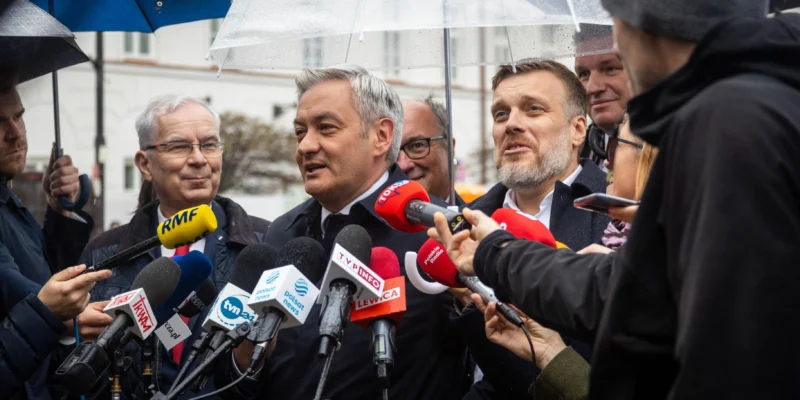 Mężczyzna w garniturze przemawia do licznych mikrofonów dziennikarzy podczas konferencji prasowej na zewnątrz, w deszczowy dzień, otoczony reporterami i parasolami.