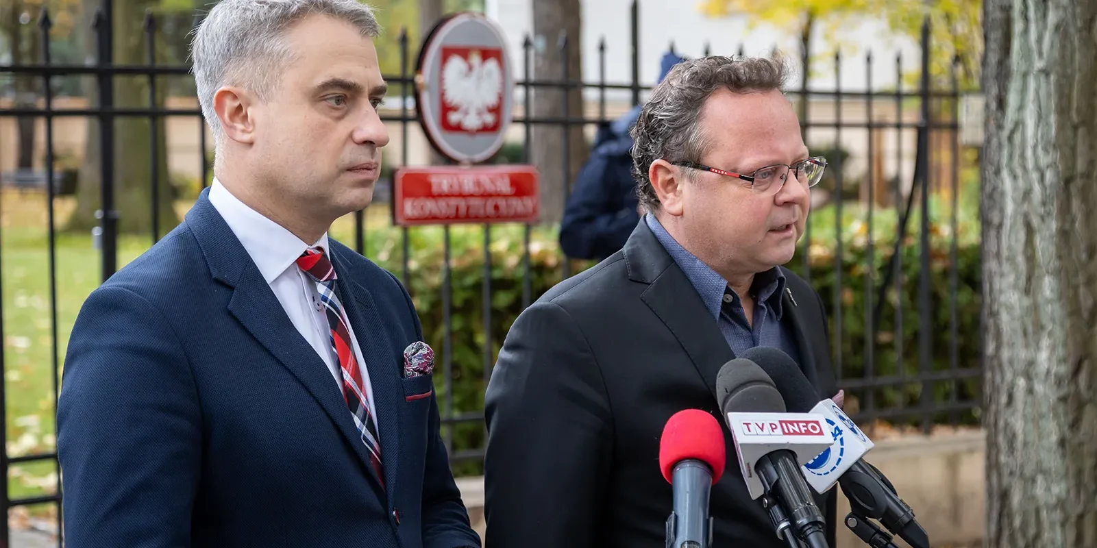 Na zdjęciu widać dwóch mężczyzn stojących przed ogrodzeniem, jeden w eleganckim garniturze z krawatem, a drugi w czarnej marynarce, obaj przygotowani do udzielania wypowiedzi przed mikrofonami.