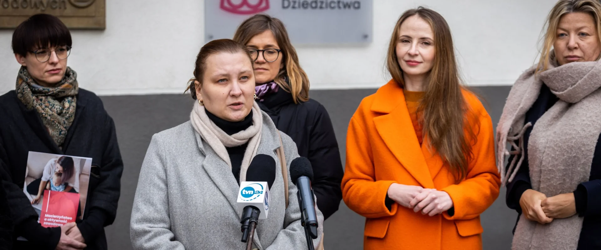 Na zdjęciu widoczne są kobiety podczas konferencji prasowej przed siedzibą Narodowego Instytutu Dziedzictwa, gdzie jedna z nich przemawia do mikrofonu.