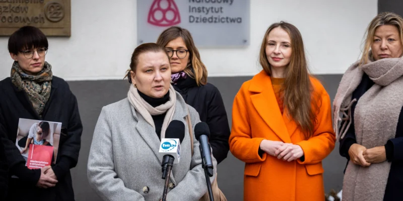 Na zdjęciu widoczne są kobiety podczas konferencji prasowej przed siedzibą Narodowego Instytutu Dziedzictwa, gdzie jedna z nich przemawia do mikrofonu.