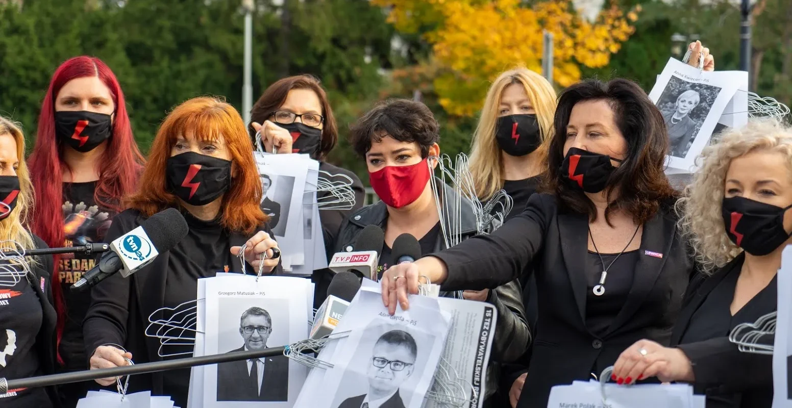 Grupa kobiet na demonstracji w parku, ubranych na czarno i noszących czarne maseczki z czerwonym symbolem błyskawicy, trzyma plakaty z wizerunkami osób i rozmawia z mediami przy mikrofonach.