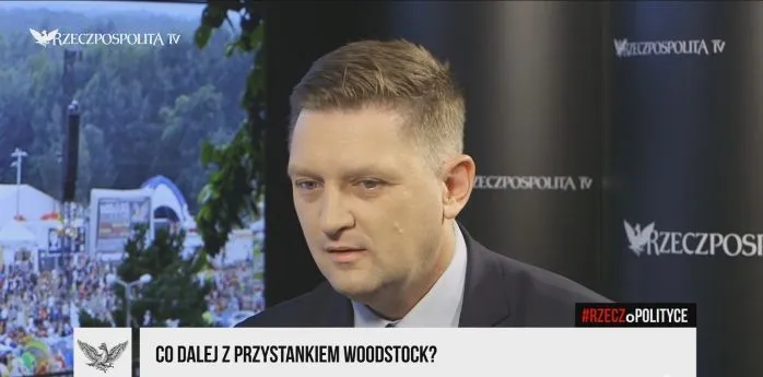 Mężczyzna w garniturze udziela wywiadu w studiu telewizyjnym; na ekranie widoczna jest plansza z pytaniem "Co dalej z Przystankiem Woodstock?" oraz zdjęcie tłumu w tle.