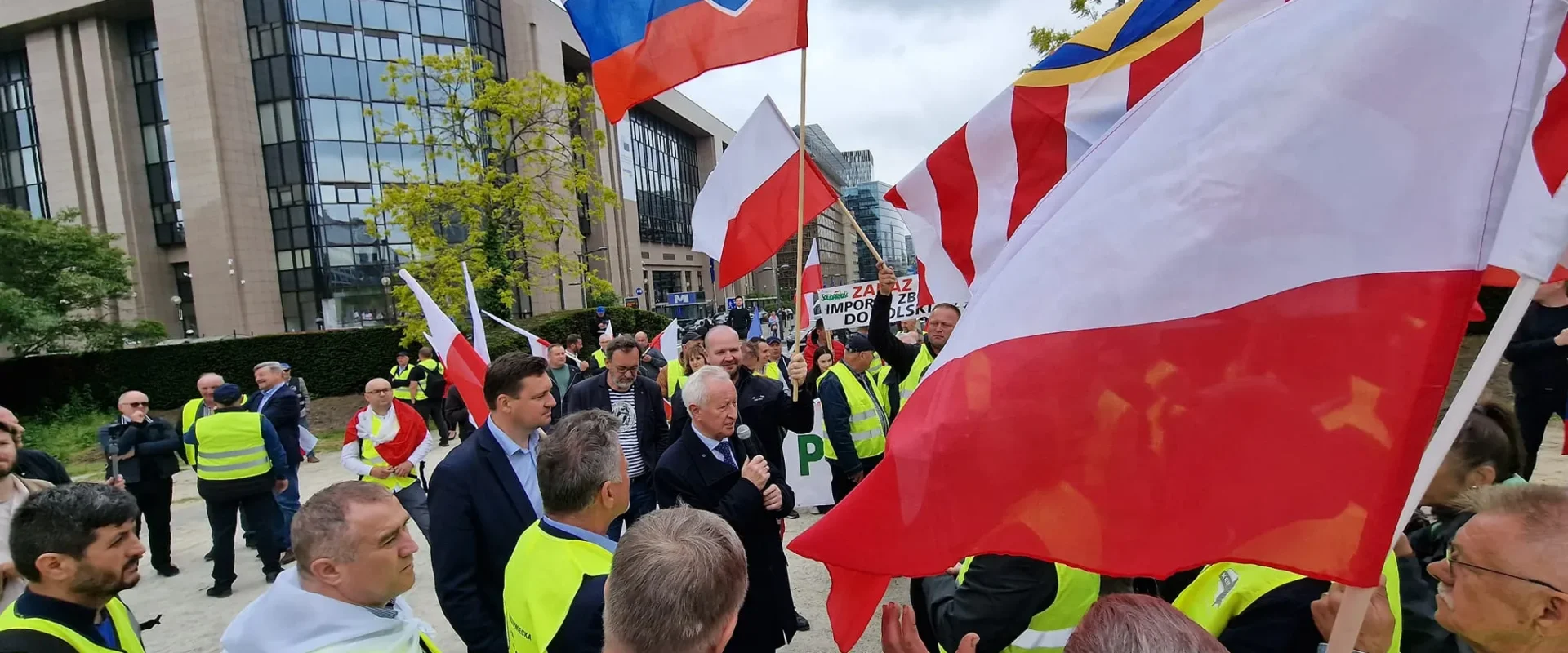 Na zdjęciu widoczna jest demonstracja z udziałem osób w kamizelkach odblaskowych, trzymających flagi Polski i Słowacji, odbywająca się w miejskim otoczeniu.