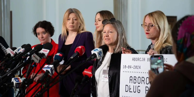 Grupa kobiet podczas konferencji prasowej w Sejmie stoi przed szeregiem mikrofonów, trzymając plakat dotyczący aborcji.