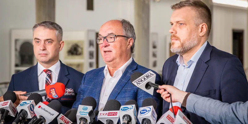 Na zdjęciu widać trzech mężczyzn z mikrofonami w tle, podczas konferencji prasowej.