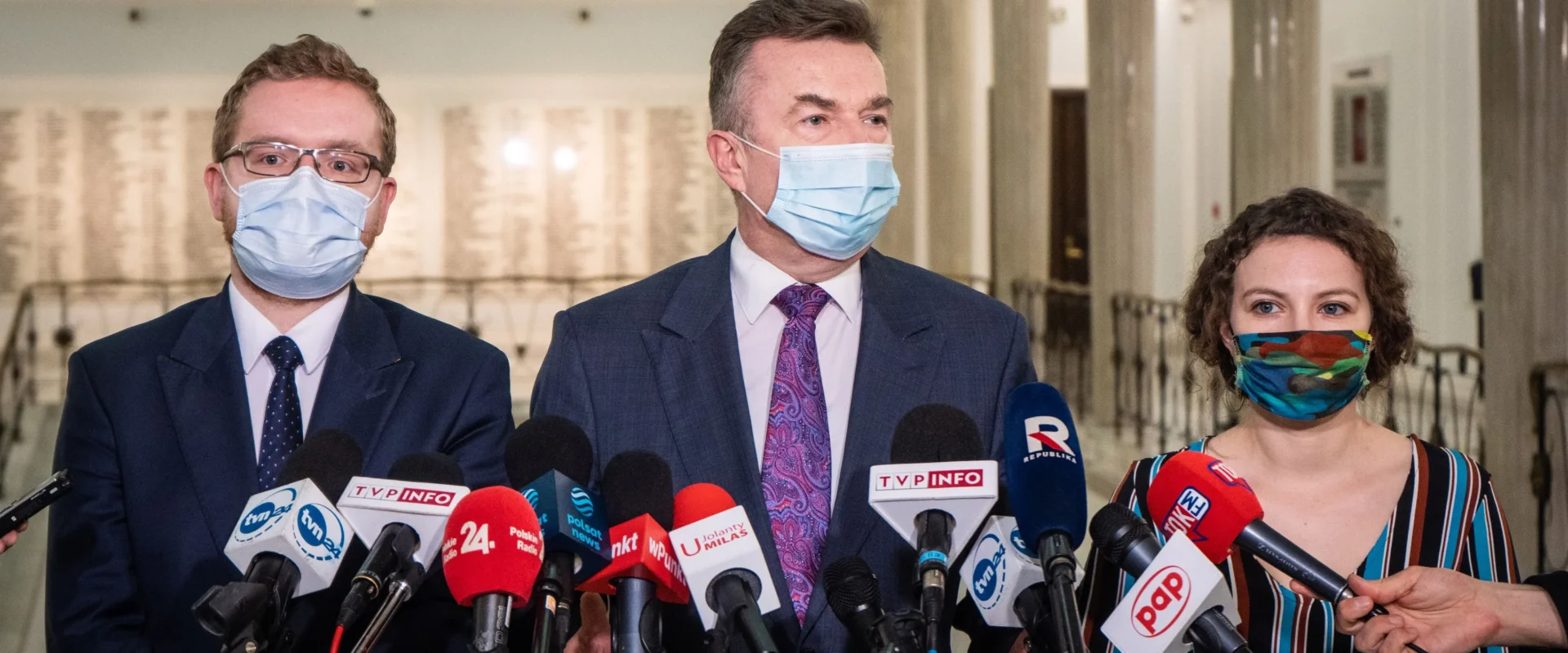 Na zdjęciu trzej mówiący do mediów politycy w maseczkach, otoczeni mikrofonami w jasnym, publicznym wnętrzu.