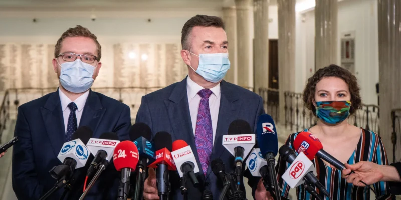 Na zdjęciu trzej mówiący do mediów politycy w maseczkach, otoczeni mikrofonami w jasnym, publicznym wnętrzu.