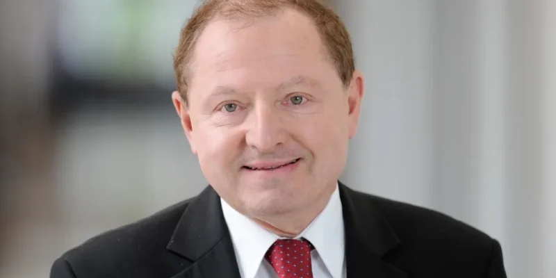 Mężczyzna w garniturze i czerwonym krawacie patrzy prosto w obiektyw i uśmiecha się.