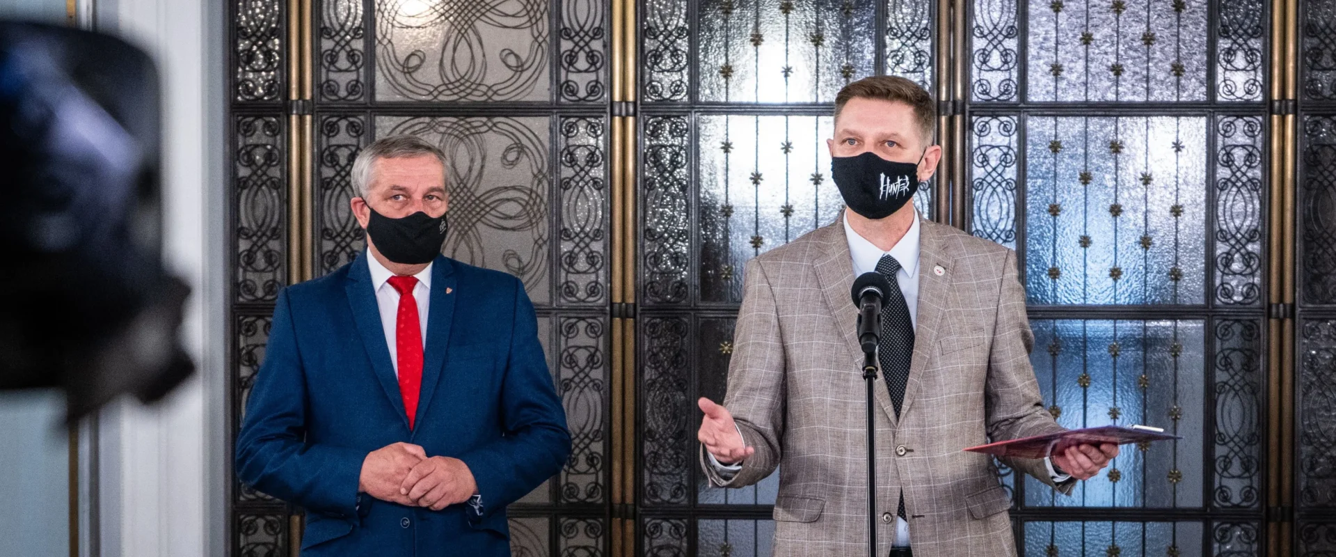Dwóch mężczyzn w garniturach i maseczkach stoi przy mikrofonie podczas konferencji prasowej, za nimi ozdobne metalowe drzwi z ornamentami.