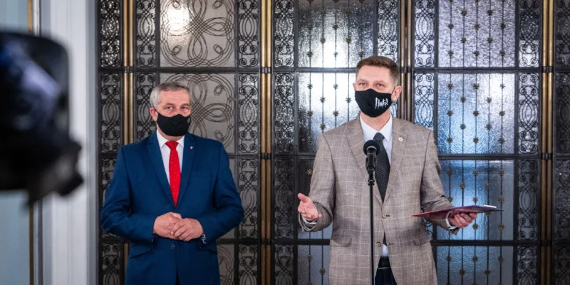 Dwóch mężczyzn w garniturach i maseczkach stoi przy mikrofonie podczas konferencji prasowej, za nimi ozdobne metalowe drzwi z ornamentami.