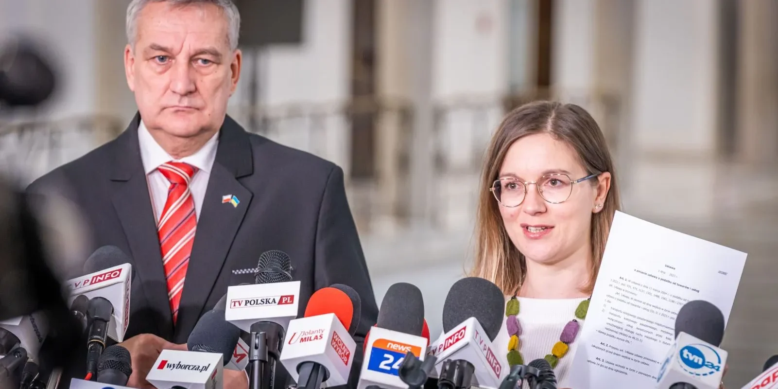 Dwóch polityków podczas konferencji prasowej otoczonej licznymi mikrofonami, omawiających obniżki cen biletów PKP związane z zerowym VAT na połączenia kolejowe.