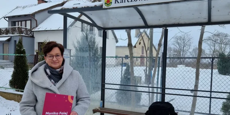 Kobieta w szarym płaszczu trzyma czerwony plakat w przystanku autobusowym w miejscowości Karczowiska Górne, otoczona śnieżnym krajobrazem.