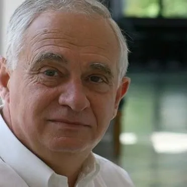 Portret starszego mężczyzny o siwych włosach i łagodnym wyrazie twarzy w białej koszuli na rozmytym, zielonkawym tle, ilustrujący artykuł o coraz większym oddawaniu władzy służbom.