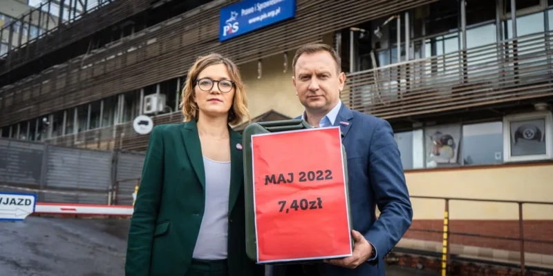 Dwóch ludzi stoi przed budynkiem z logo PiS i trzyma duży czerwony karton z napisem MAJ 2022 7,40 zł.