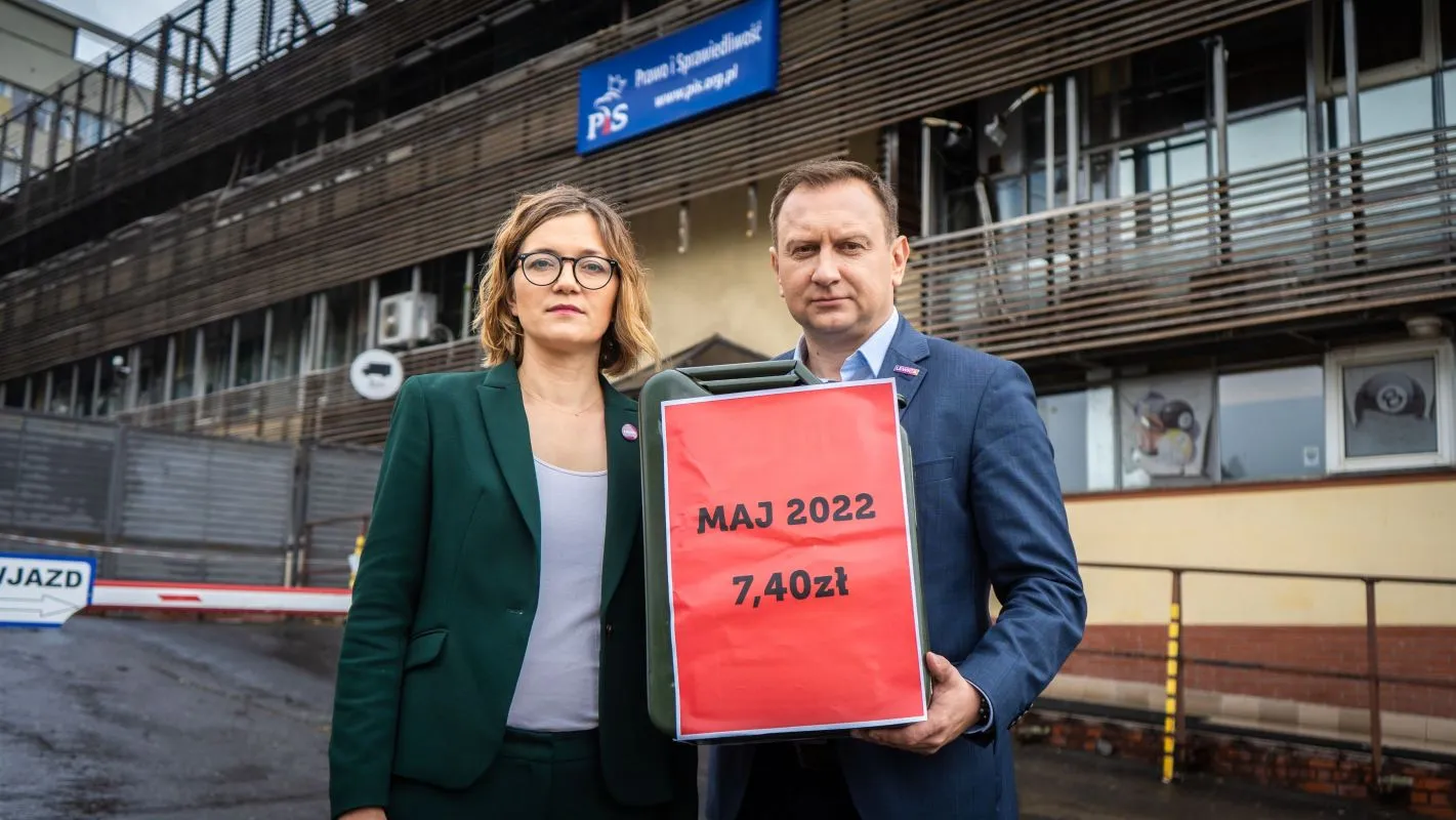 Dwóch ludzi stoi przed budynkiem z logo PiS i trzyma duży czerwony karton z napisem MAJ 2022 7,40 zł.