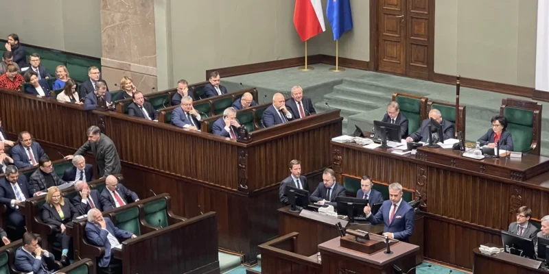 Na zdjęciu widoczna jest sala sejmowa, w której posłowie dyskutują na temat braku funduszy z KPO, a w centralnej części przemawia mężczyzna.