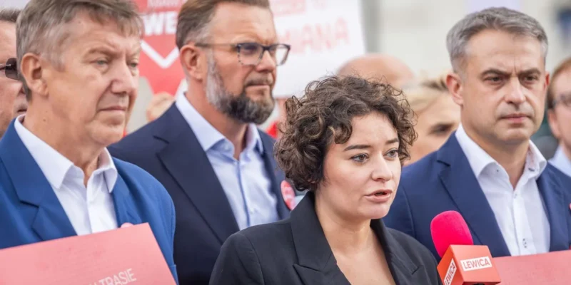 Kobieta w czarnym garniturze przemawia do mikrofonu z logo Lewicy podczas konferencji, otoczona przez mężczyzn w garniturach i czerwone banery w tle.