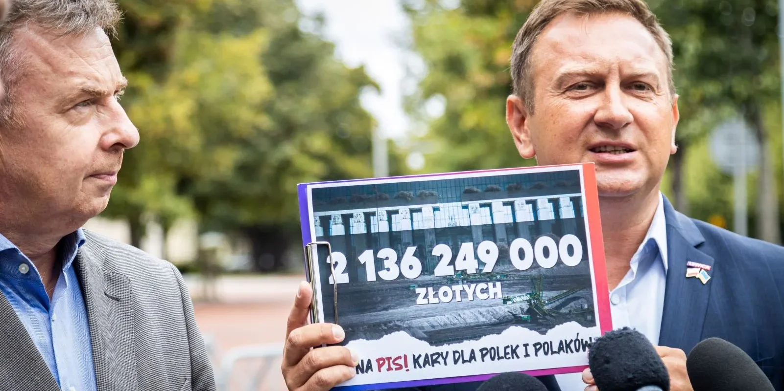 Na zdjęciu dwóch mężczyzn podczas konferencji prasowej, gdzie jeden z nich trzyma kartkę z informacją o kwocie 2 136 249 000 złotych oraz przesłaniu krytykującym rząd.