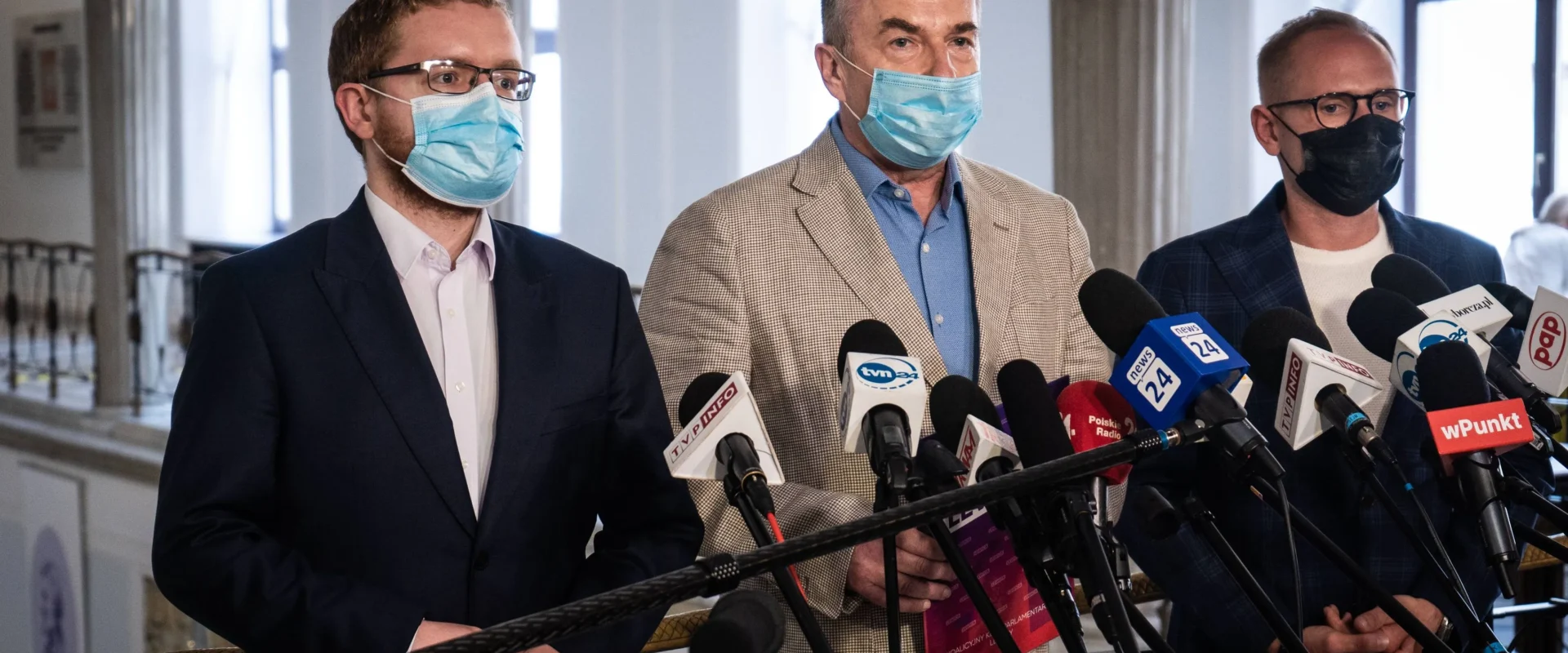 Trzy osoby w garniturach i maseczkach stojące za mikrofonami podczas konferencji prasowej.