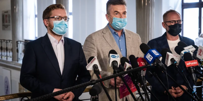 Trzy osoby w garniturach i maseczkach stojące za mikrofonami podczas konferencji prasowej.