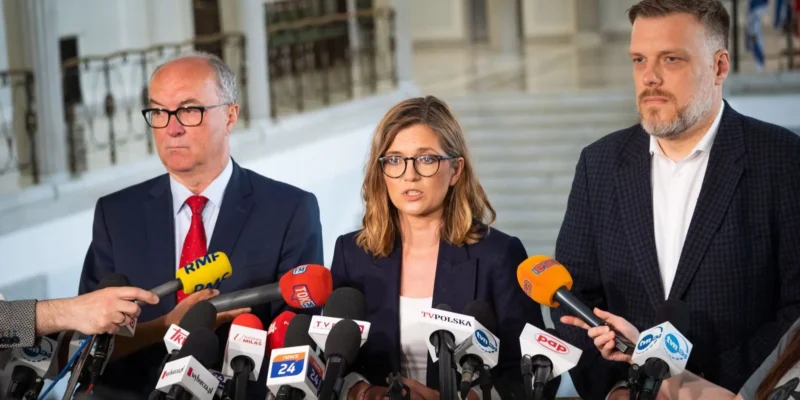 Kobieta w okularach przemawia do licznych mikrofonów podczas konferencji prasowej, stojąc między dwoma mężczyznami, w tle widoczna sala i logotypy mediów, związana z komisją ds. badania wpływów rosyjskich.