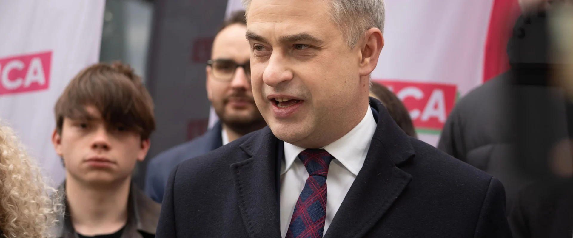 Mężczyzna w garniturze przemawia do mediów podczas konferencji prasowej, w tle widoczne banery Lewicy.