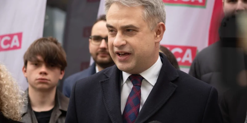 Mężczyzna w garniturze przemawia do mediów podczas konferencji prasowej, w tle widoczne banery Lewicy.