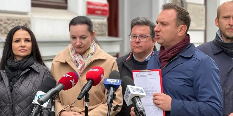 Grupa osób na konferencji prasowej na zewnątrz budynku; mężczyzna z teczką przemawia do zestawu mikrofonów, otoczony przez dwie kobiety i innych mężczyzn.