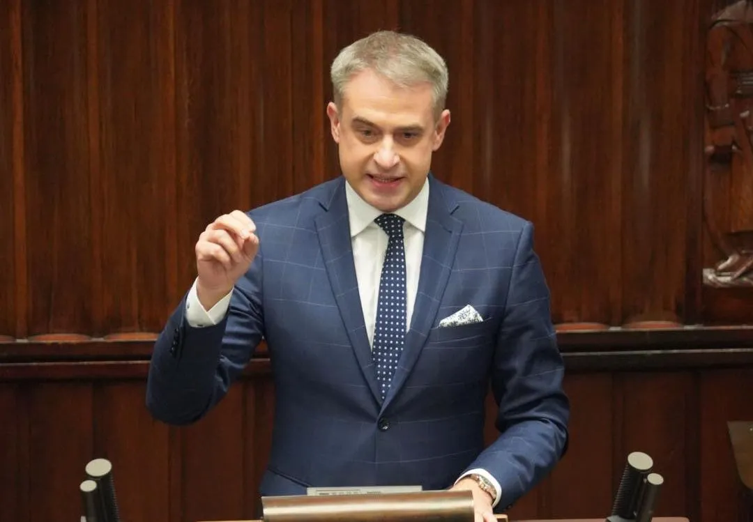 Mężczyzna w niebieskim garniturze wygłasza przemówienie w parlamencie przed drewnianym tłem.