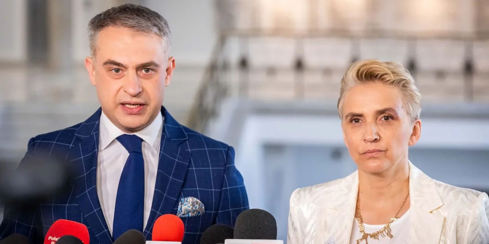 Dwóch polityków podczas konferencji prasowej stoi przed mikrofonami; mężczyzna w granatowym garniturze w kratę i niebieskim krawacie, kobieta w białej marynarce z naszyjnikiem.