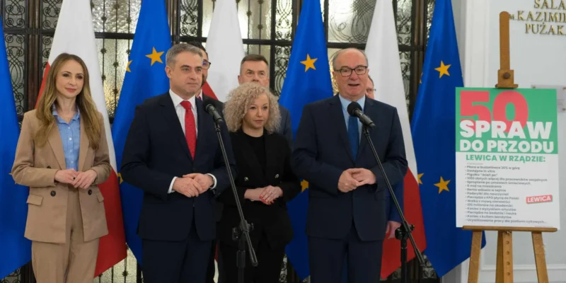 Grupa polityków stoi przy mikrofonach podczas konferencji prasowej, na tle flag Polski i Unii, a obok widnieje plakat „50 Spraw do Przodu” promujący Lewicę w rządzie.