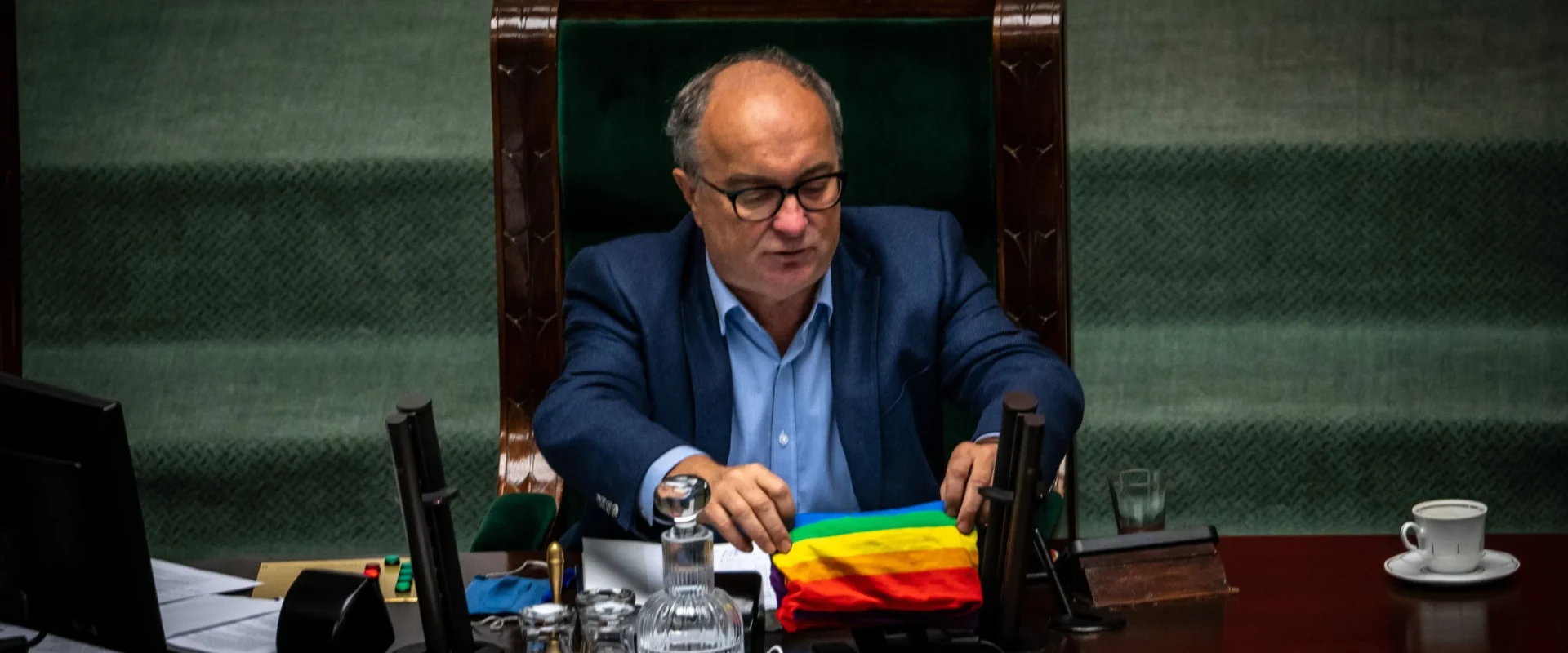 Na zdjęciu widać mężczyznę w okularach siedzącego przy biurku w parlamencie z tęczową flagą przed sobą.