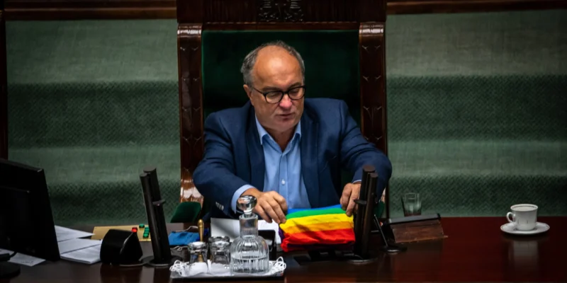 Na zdjęciu widać mężczyznę w okularach siedzącego przy biurku w parlamencie z tęczową flagą przed sobą.